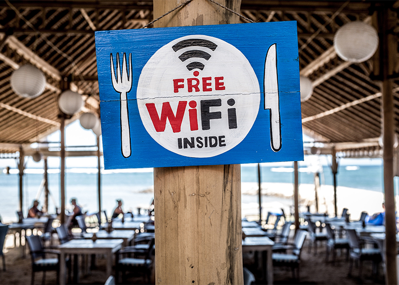 A flyer with a blue background, a white fork on the left, a white knife on the right. In the center of the poster is a white circle with black and red text that reads "Free WiFi inside." The sign is attached to a wooden beam, in the foreground, at a restaurant filled with tables and chairs.