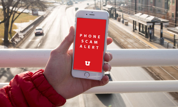 A person, who's standing on a bridge over a four-lane street, holds a phone. On its screen, it says "Phone scam alert" in white text with a white block U at the bottom, on top of a red background.